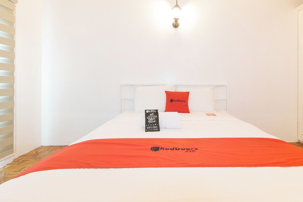 A hotel room with a bed, a red pillow, a white headboard, a red blanket, and a RedDoorz branded pillow.