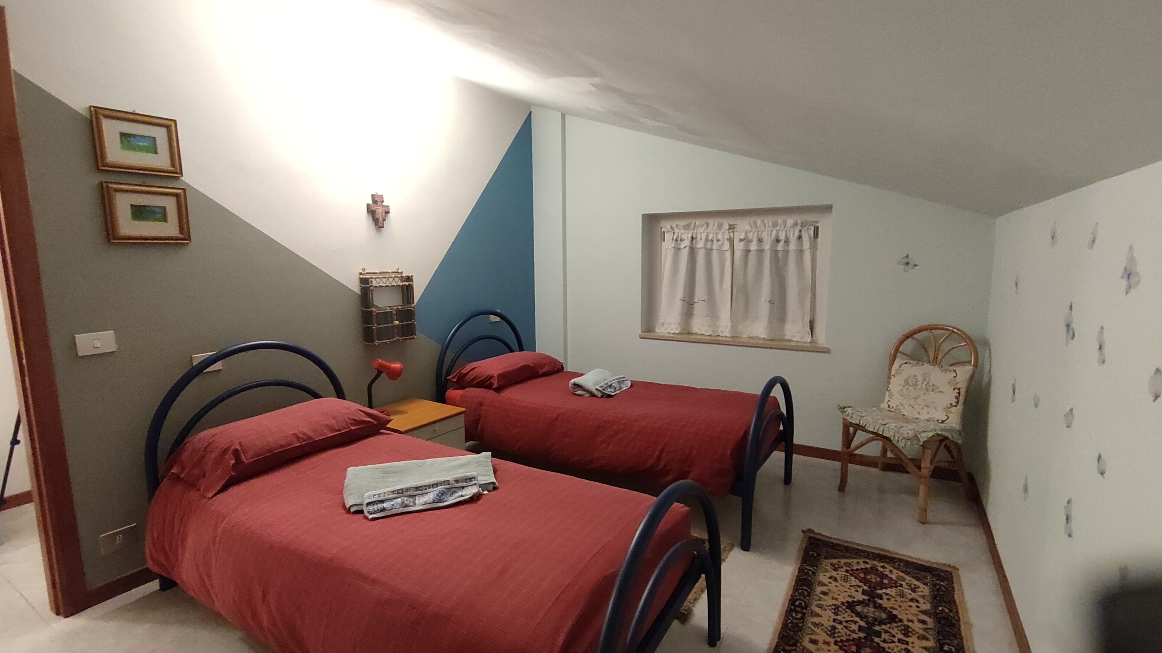 A bedroom with two single beds, a small table, a chair, a window with curtains, and a wall-mounted shelf.