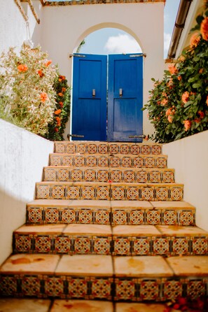 Um conjunto de degraus com azulejos decorados leva a uma porta azul, rodeada por plantas floridas.