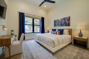 Una camera da letto con un letto, una scrivania, una sedia, una finestra con tende e un ventilatore a soffitto, oltre a un quadro appeso al muro.