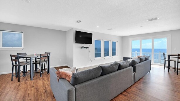 A modern living room with a grey sofa, wooden flooring, a dining area with a table and chairs, a flat-screen TV mounted on the wall, and large windows offering a view of the sea. - Oceanfront 9BR Ponte Vedra Estate with Private Beach Access. Sleeps 20! (Ponte Vedra)