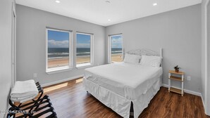 A bedroom with a large bed, two windows with a beach view, wooden flooring, and a small bedside table with a plant. - Oceanfront 9BR Ponte Vedra Estate with Private Beach Access. Sleeps 20! (Ponte Vedra)