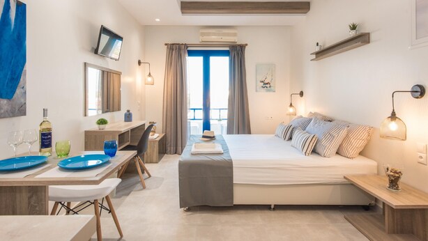 A modern bedroom with a bed, bedside tables, a desk, and a chair. There is a window with curtains, a TV mounted on the wall, and a small plant on the desk. - Mikes Beach Lux (Hersonissos)