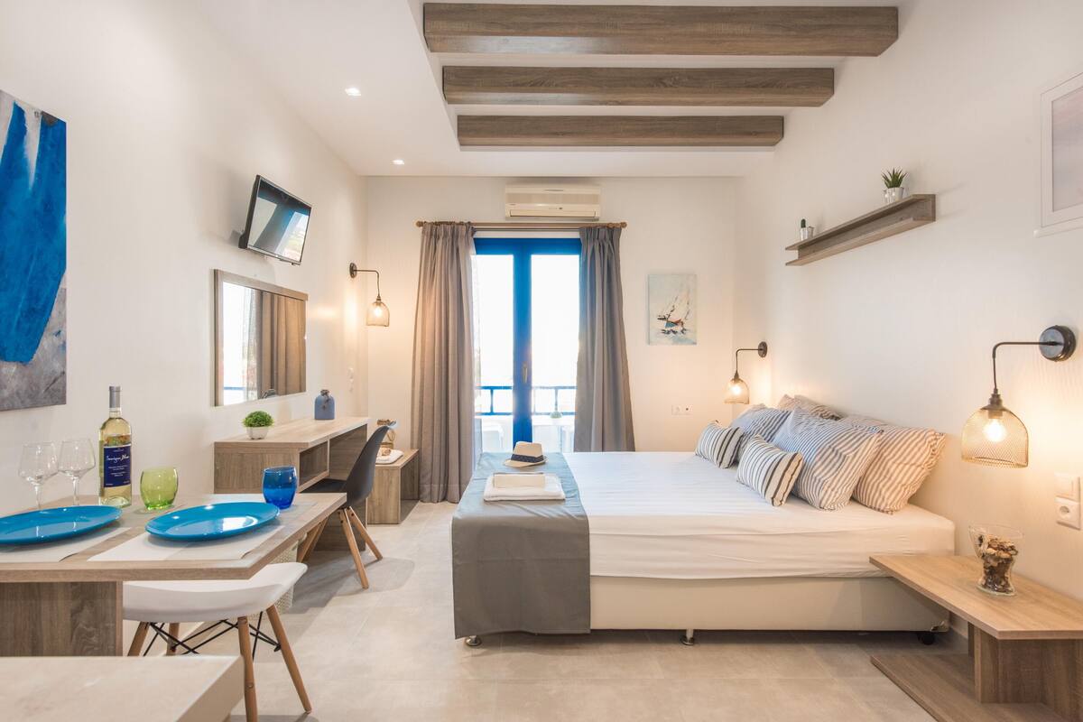 Ein modernes Schlafzimmer mit Bett, Nachttischen, Schreibtisch und Stuhl. Es gibt ein Fenster mit Vorhängen, einen an der Wand montierten Fernseher und eine kleine Pflanze auf dem Schreibtisch.