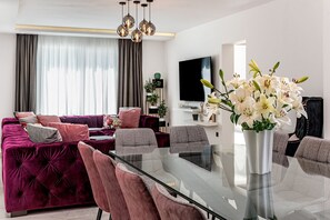 A modern living room with a glass dining table, plush seating, a flat-screen TV, and a vase of white flowers.