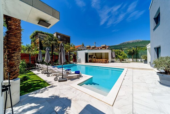 A modern pool area with lounge chairs, a barbecue area, and a view of a mountain.