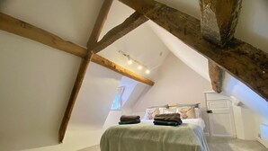 A bedroom with a sloped ceiling, exposed wooden beams, a bed with a green bedspread, and a window with curtains. - Beautiful 17th Century Coach House (Marlborough)