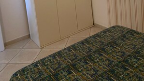 Ein Schlafzimmer mit großem Kleiderschrank, einem Bett mit gemustertem Überwurf und einem Fenster mit vertikalen Rollläden.