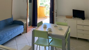 Ein kleiner, gut beleuchteter Raum mit einem blauen Sofa, einem Esstisch mit vier Stühlen, einem Fernseher auf einem Schrank und einem Balkon mit Blick ins Grüne.