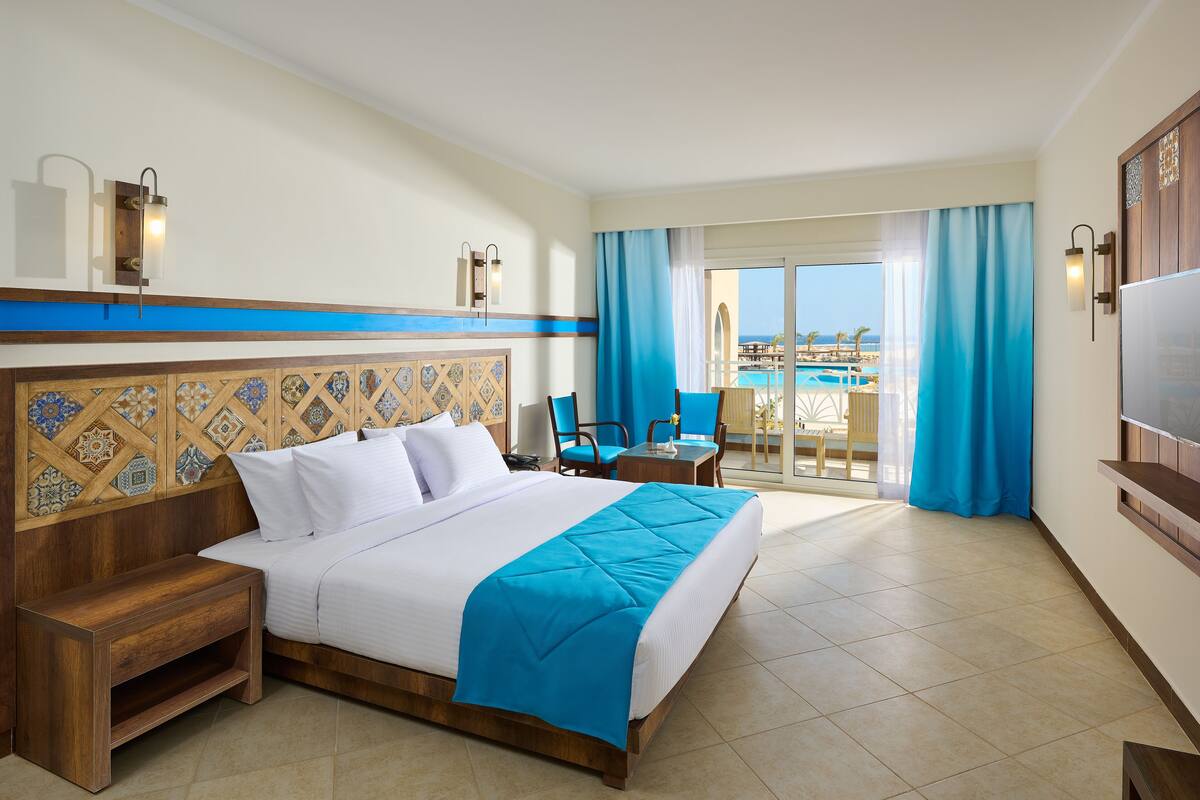 A hotel room with a bed, a wooden nightstand, a desk with a chair, and a view of the pool and ocean through large windows.