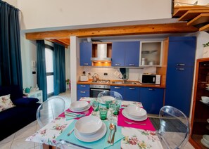Une cuisine compacte avec des meubles bleus, un plafond en bois et un coin repas agrémenté de verres et d’assiettes transparents.