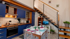 A modern kitchen with blue cabinets, a wooden staircase, and a dining area with clear chairs and a floral tablecloth.