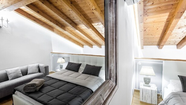 A bedroom with a wooden ceiling, a bed with a grey and black duvet, a grey sofa, and a bedside table with a lamp. - Starfish 4         . (Venezia)