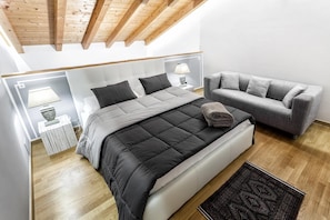 A bedroom with a bed, a grey sofa, a bedside table with a lamp, and a patterned rug.