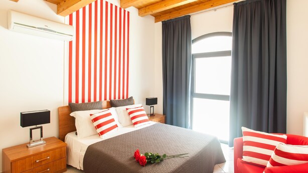 A bedroom with a bed, nightstands, a lamp, a red and white striped headboard, a dark curtain, and a red chair.