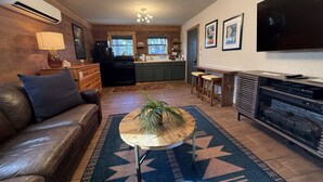 A living room with a leather sofa, a round coffee table with a plant, a fireplace, a television, and a kitchen in the background.