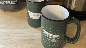 A coffee brewing station with a Mountain Bird Coffee & Tea Company kettle, two mugs labeled 'Welcome to Lobolly Pines Adventure Camp,' and a jar of Mountain Bird coffee.