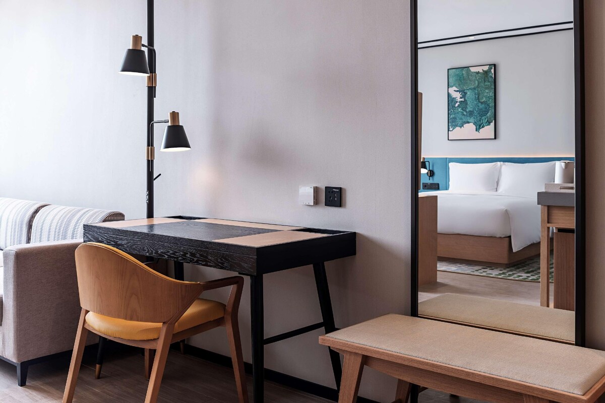 Habitación de hotel moderna con un escritorio de madera, una silla, un banco y un espejo grande que refleja una cama y un cuadro en la pared.