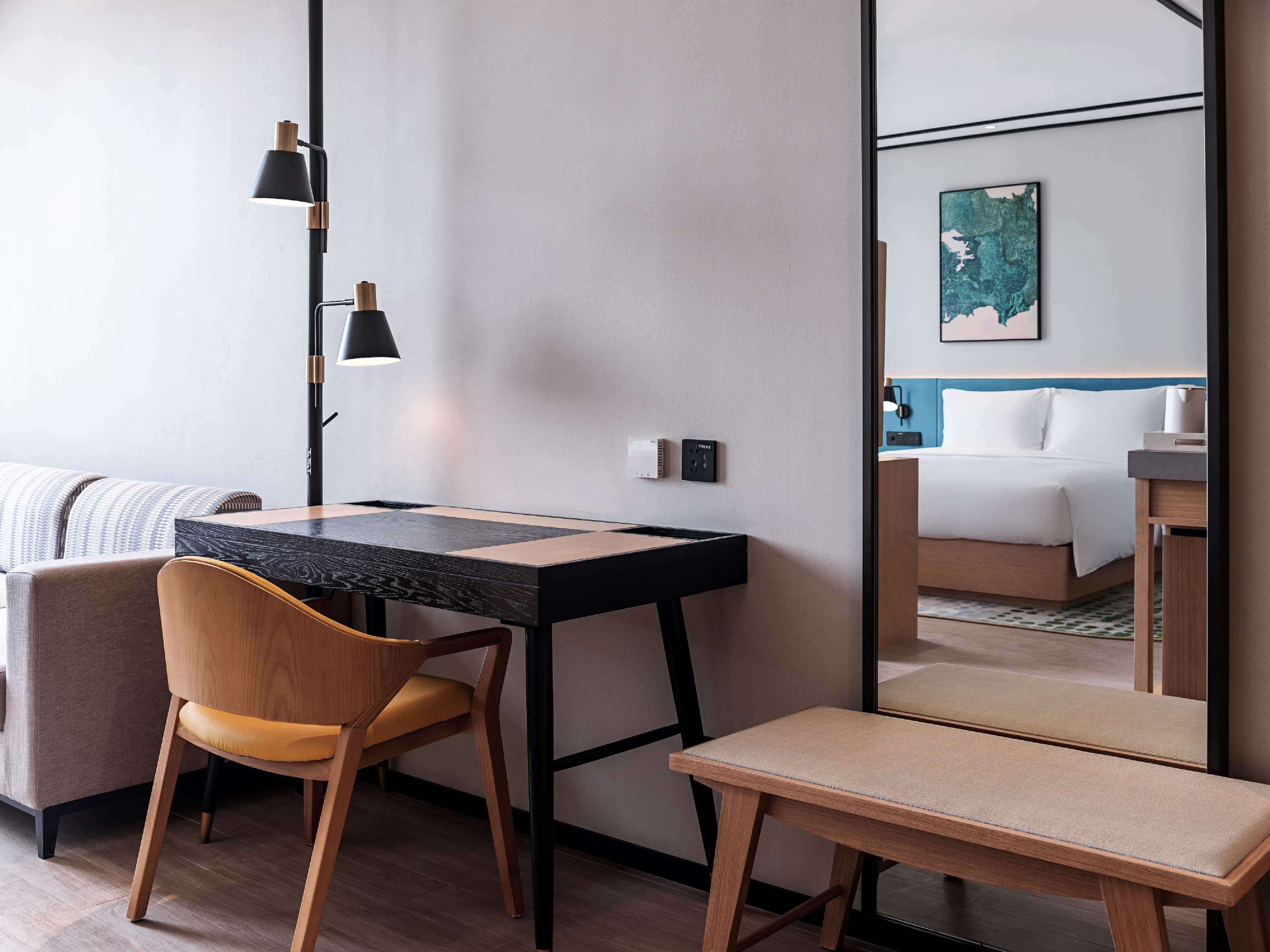 Una habitación de hotel moderna con un escritorio de madera, una silla, un banco y un espejo grande que refleja una cama y un cuadro.