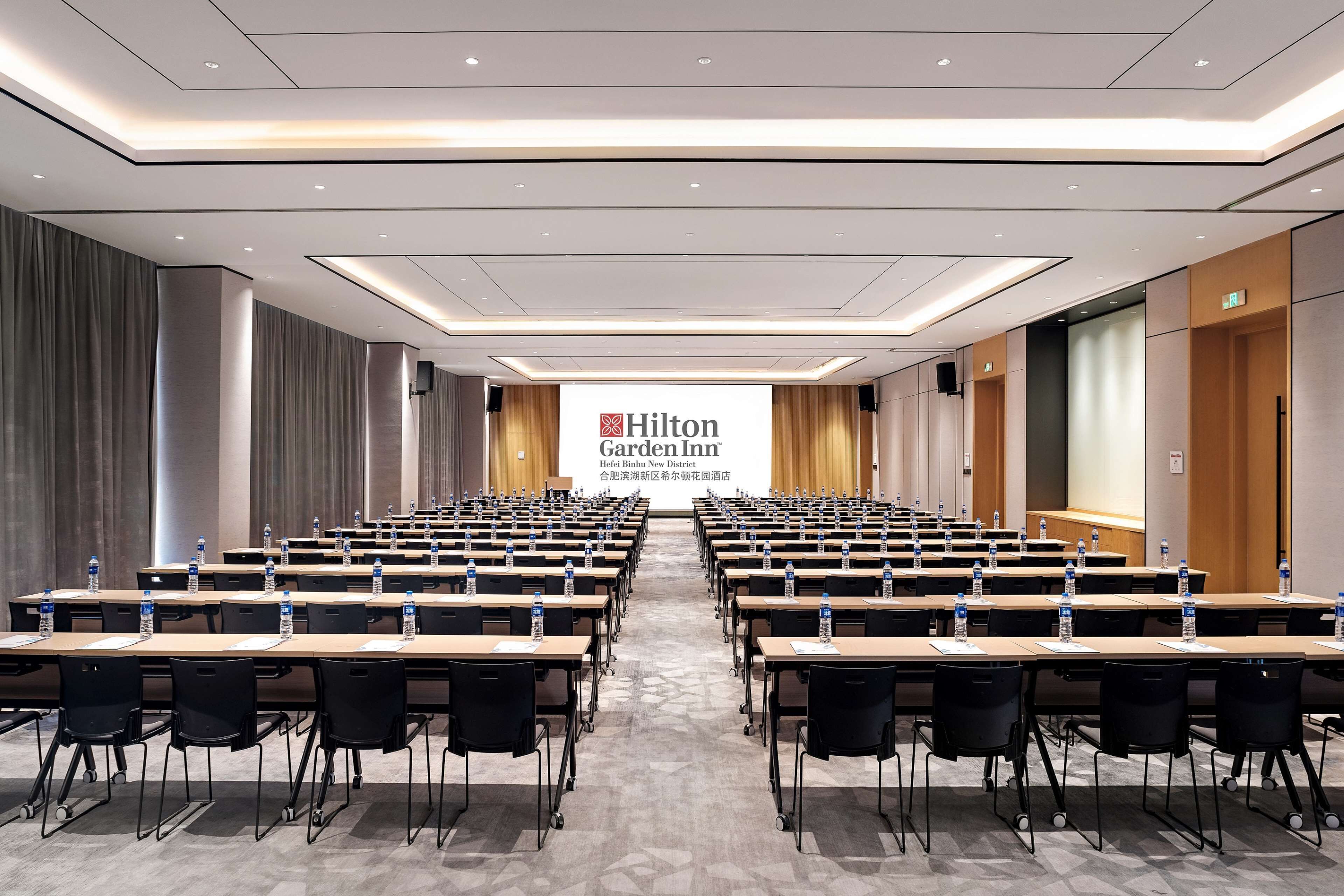 A conference room with rows of tables, chairs, and water bottles, featuring a Hilton Garden Inn sign.