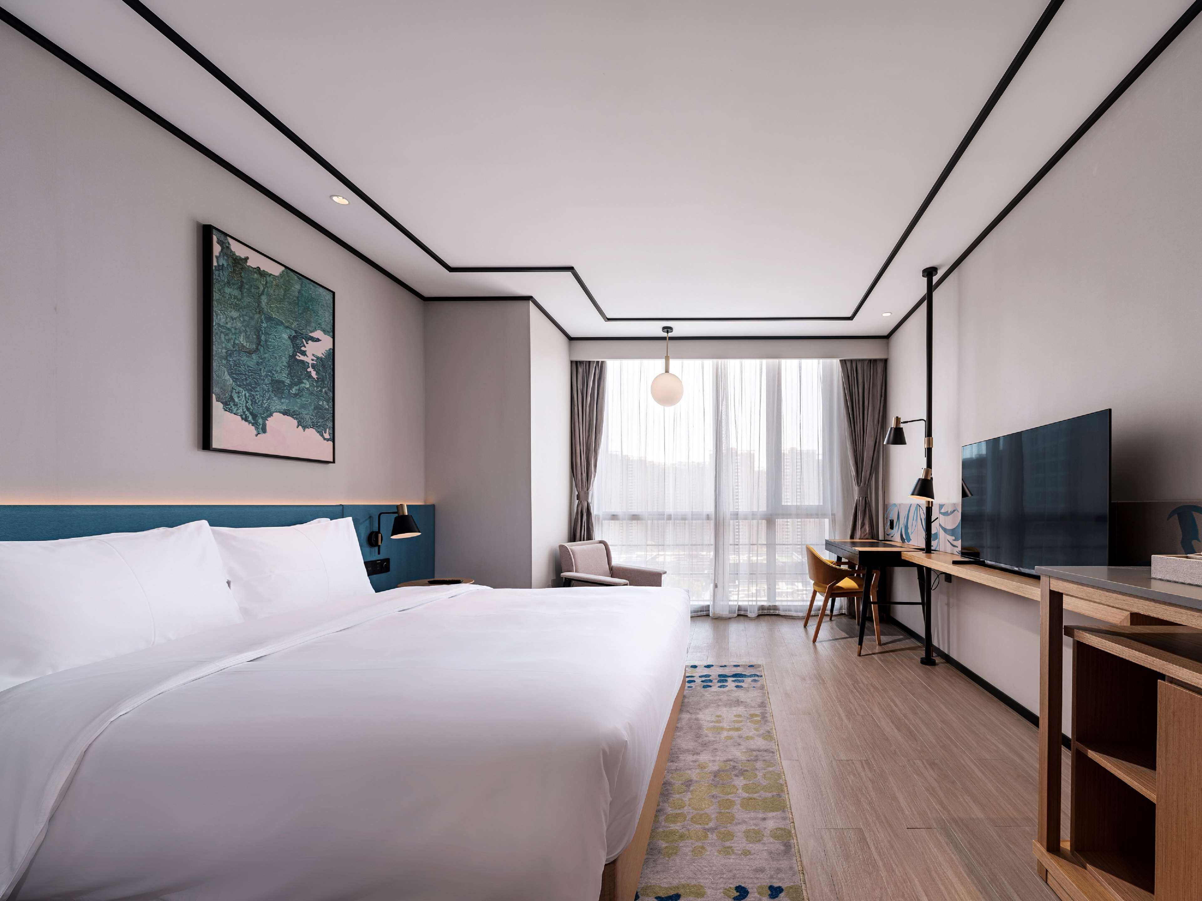 A modern hotel room with a large bed, a desk with a TV, a chair, and a view of the city through large windows.