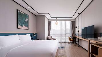 A modern hotel room with a large bed, a desk with a TV, a chair, and a view of the city through large windows.