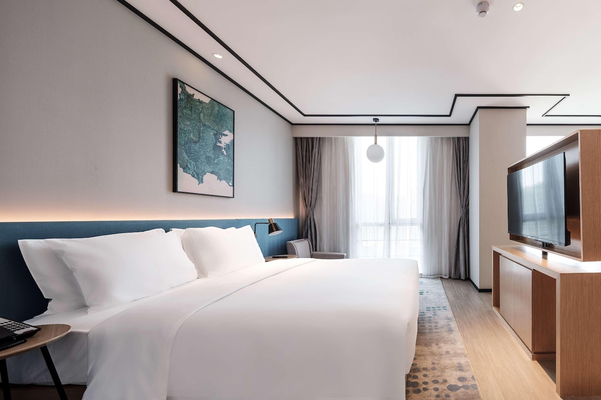 A modern hotel room with a large bed, a desk, a flat-screen TV, and a large window with curtains.