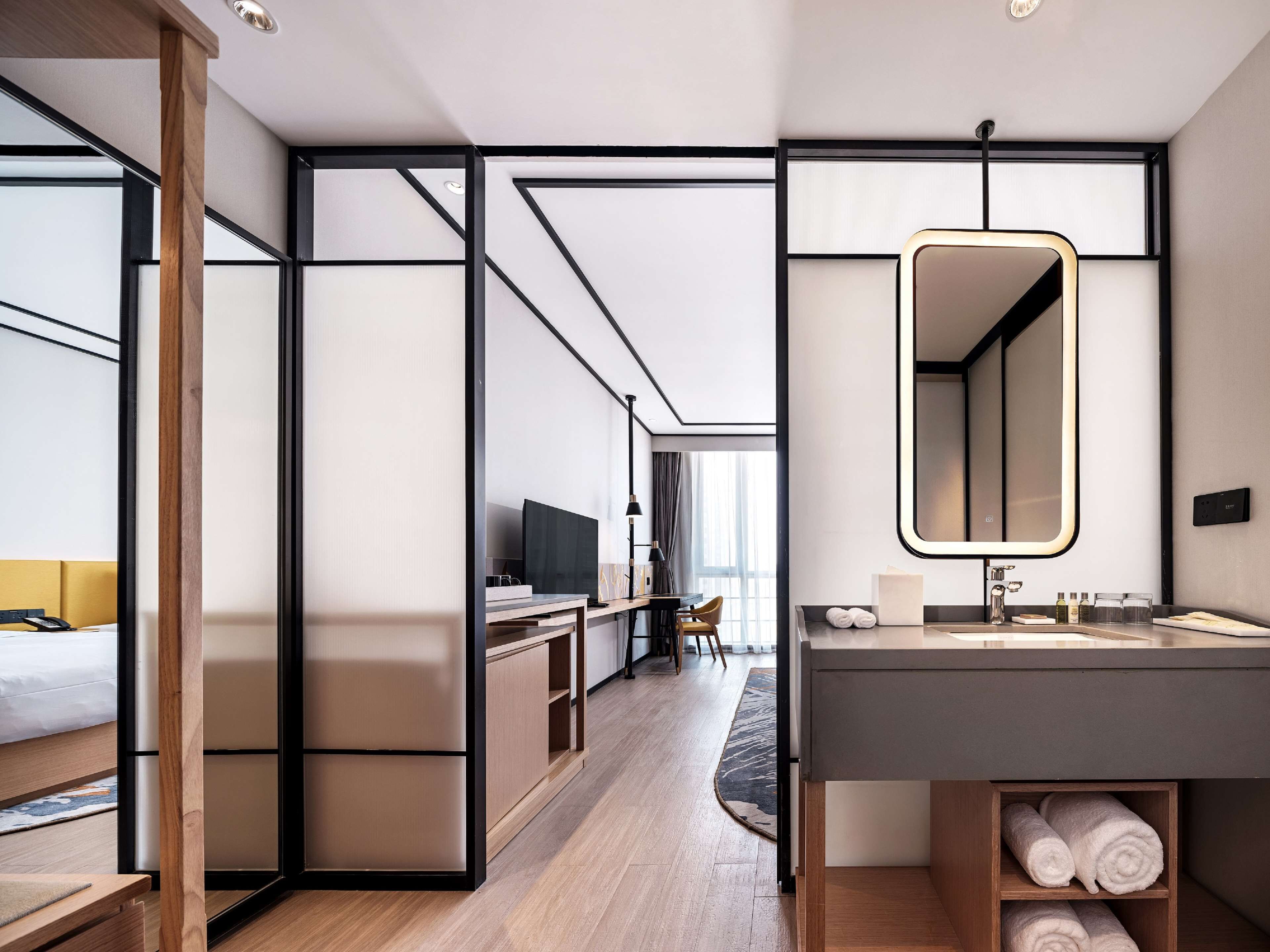 Una habitación de hotel moderna con piso de madera, un espejo grande, un escritorio con una silla, una cama con cabecera amarilla y vista a la ciudad a través de amplios ventanales.