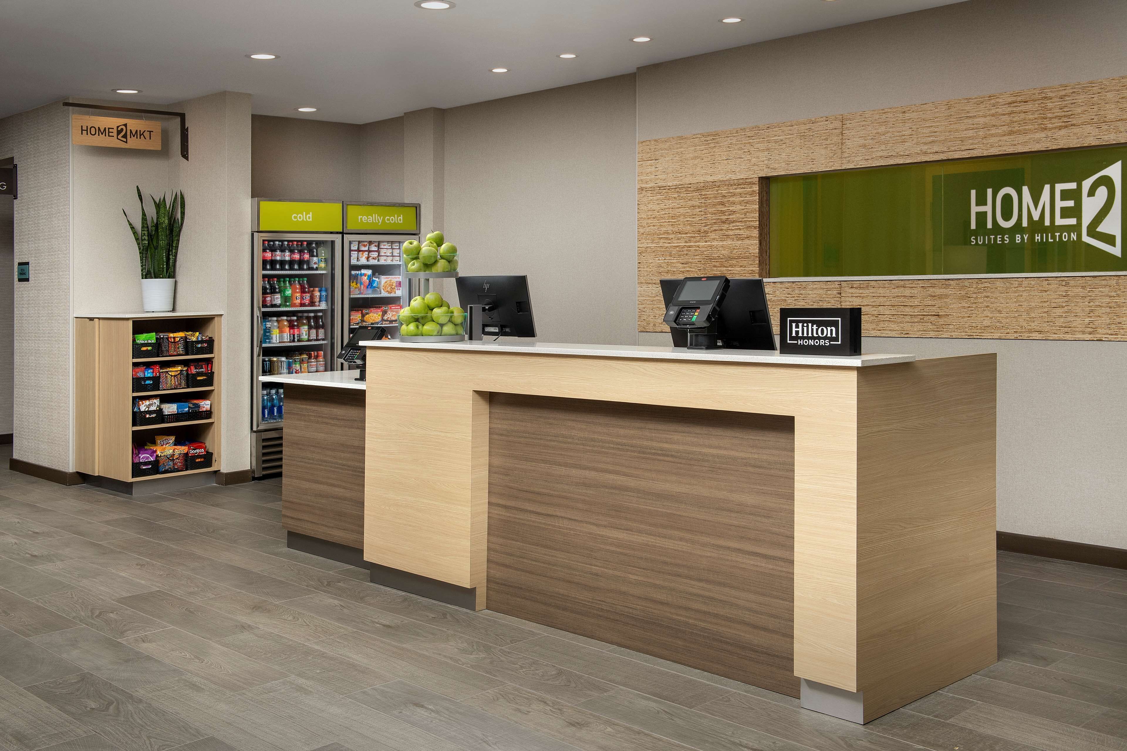 A hotel reception area with a wooden reception desk, a plant, a vending machine, and a sign for 'HOME 2 Suites by Hilton'.