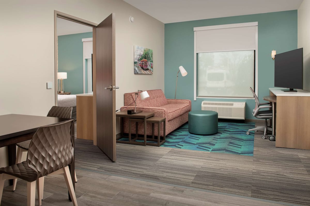 A modern hotel room with a dining area, a sofa, a TV, and a window with a view.