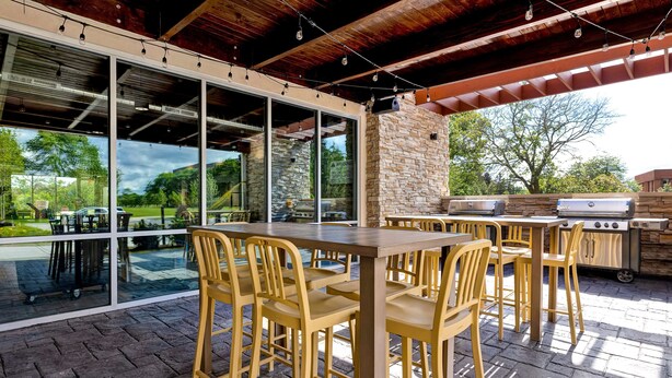 An outdoor patio with a wooden table and chairs, a stone wall, and a grill. - Home2 Suites by Hilton Lincolnshire Chicago (Lincolnshire)