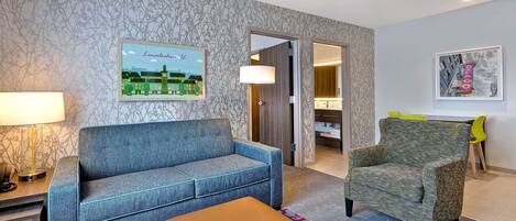 A hotel room with a blue sofa, a green armchair, a wooden coffee table, and a patterned rug.