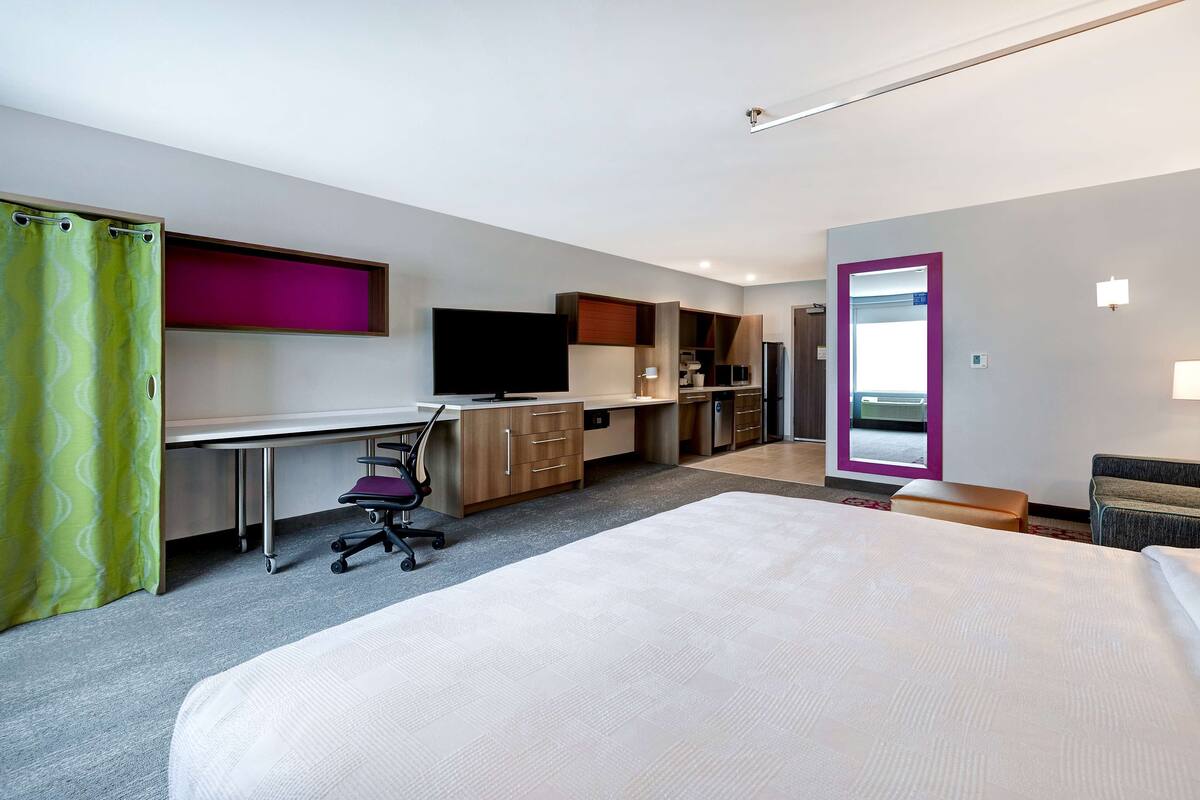 Una camera d'albergo con un letto, una scrivania, una sedia, angolo cottura e bagno con specchio.