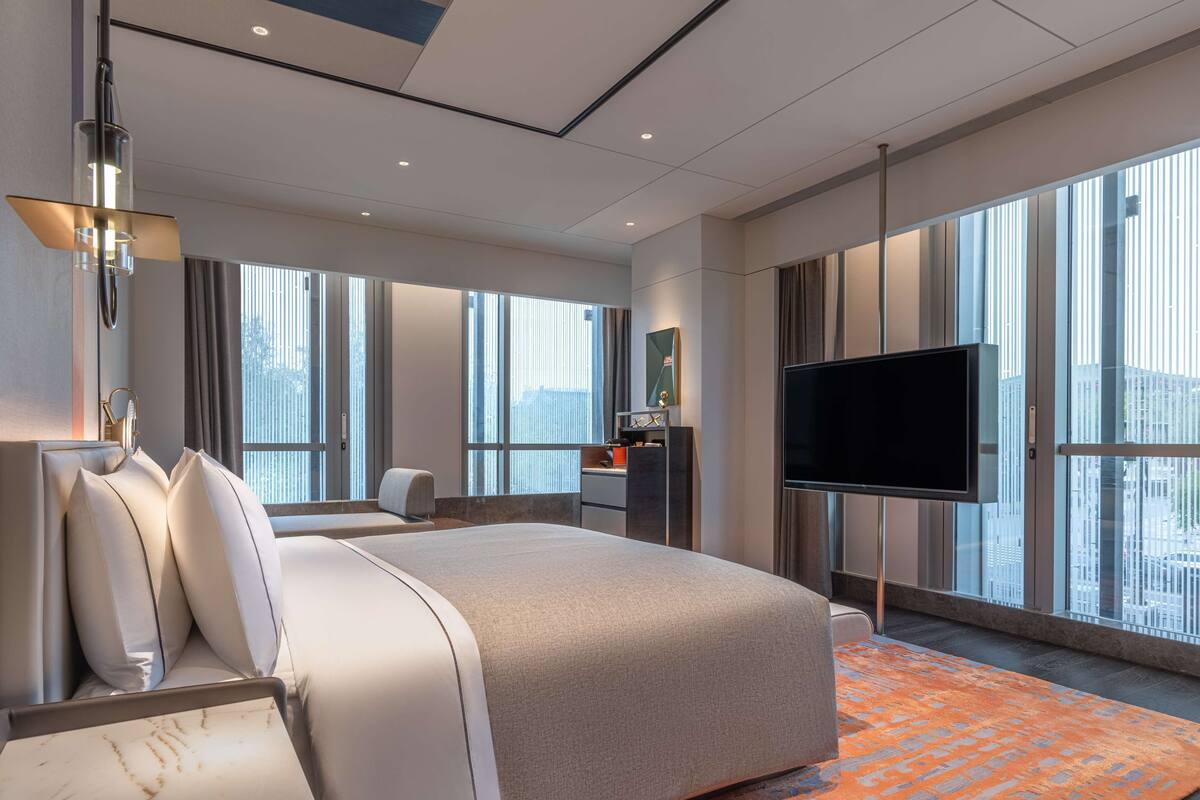 A modern hotel room with a large bed, a flat-screen TV, a desk, and a large window offering a city view.