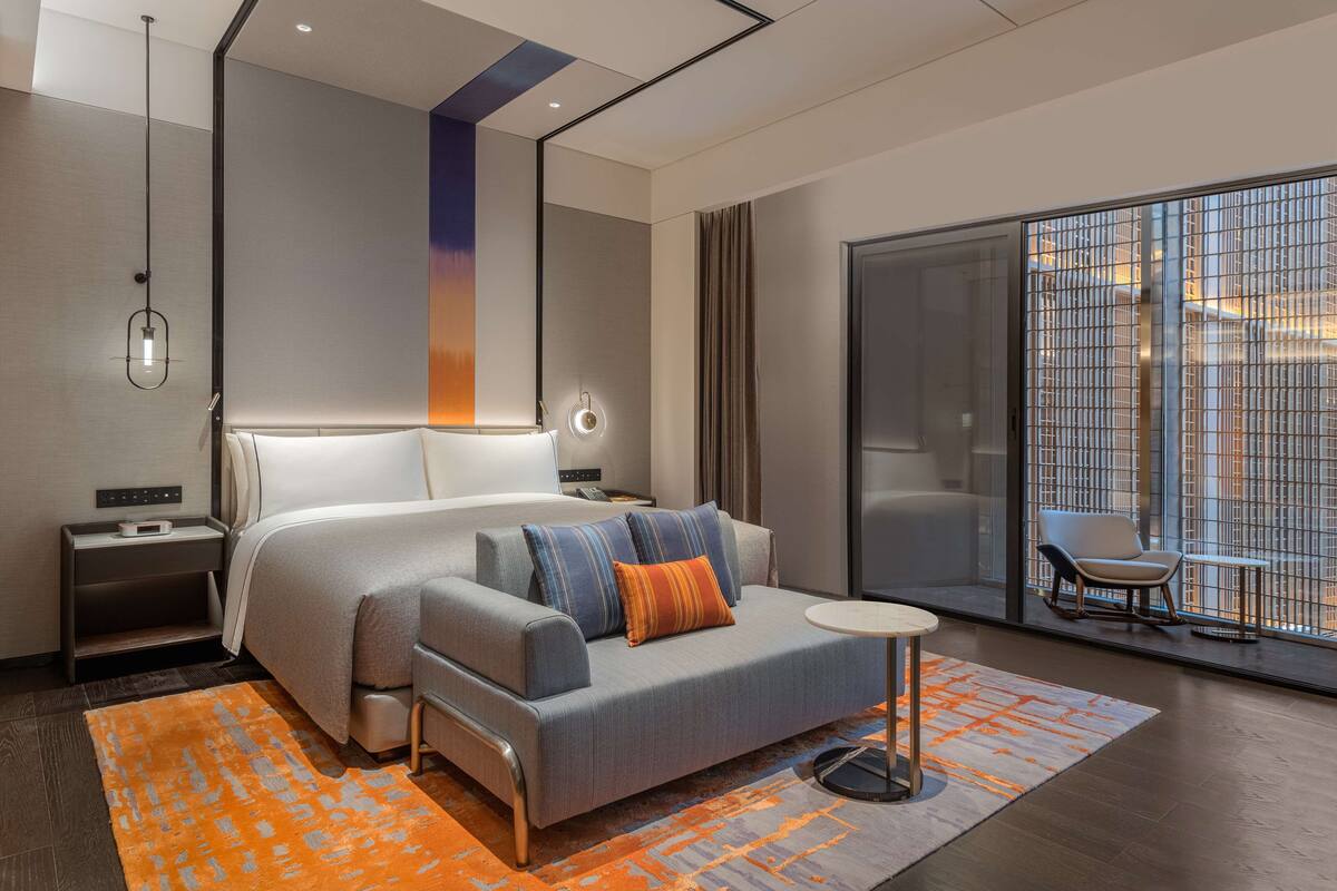 A modern hotel room with a large bed, a sofa, a small table, and a view of the city through a large window.