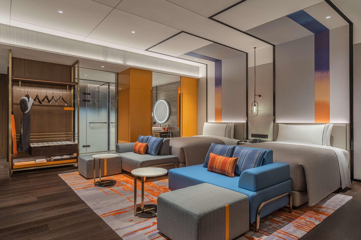 A modern hotel room with a large bed, two sofas, a small table, and a walk-in closet with a mirror.