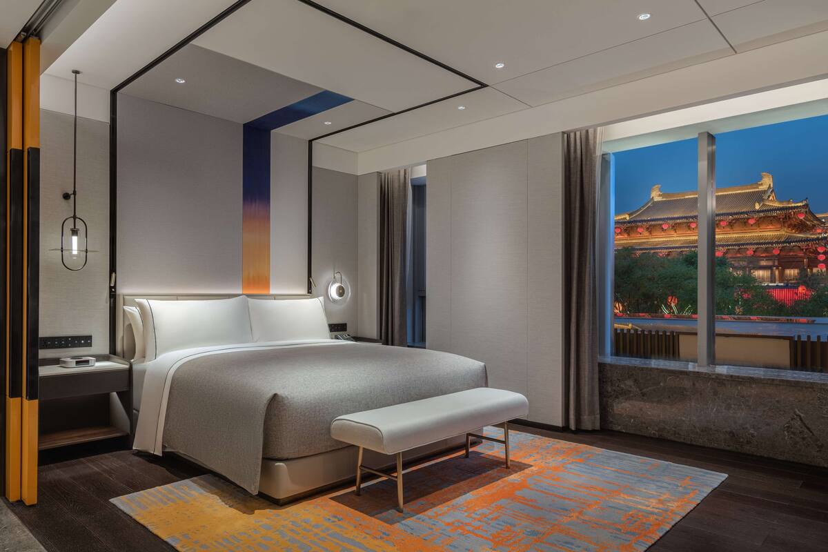 A modern hotel room with a large bed, bedside tables, a bench, and a view of traditional architecture through a window.