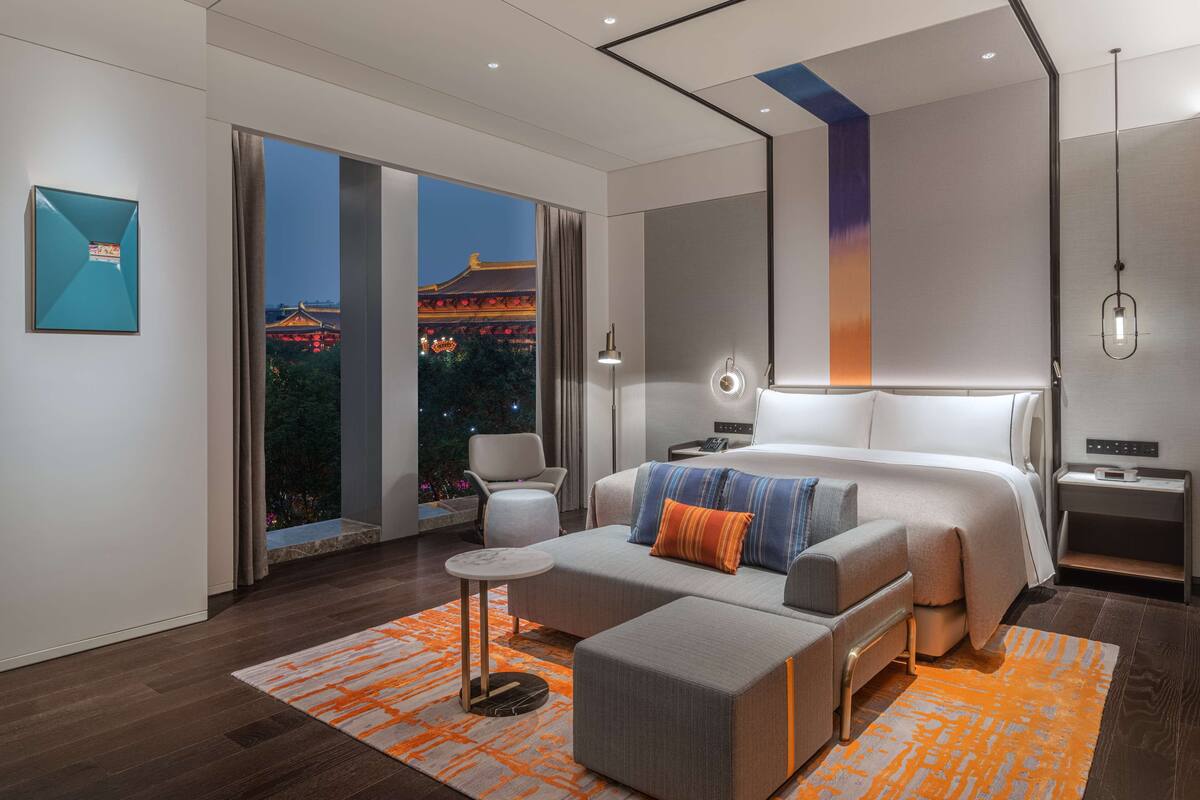 A modern hotel room with a large bed, a sofa, a small table, and a view of a traditional building at night.