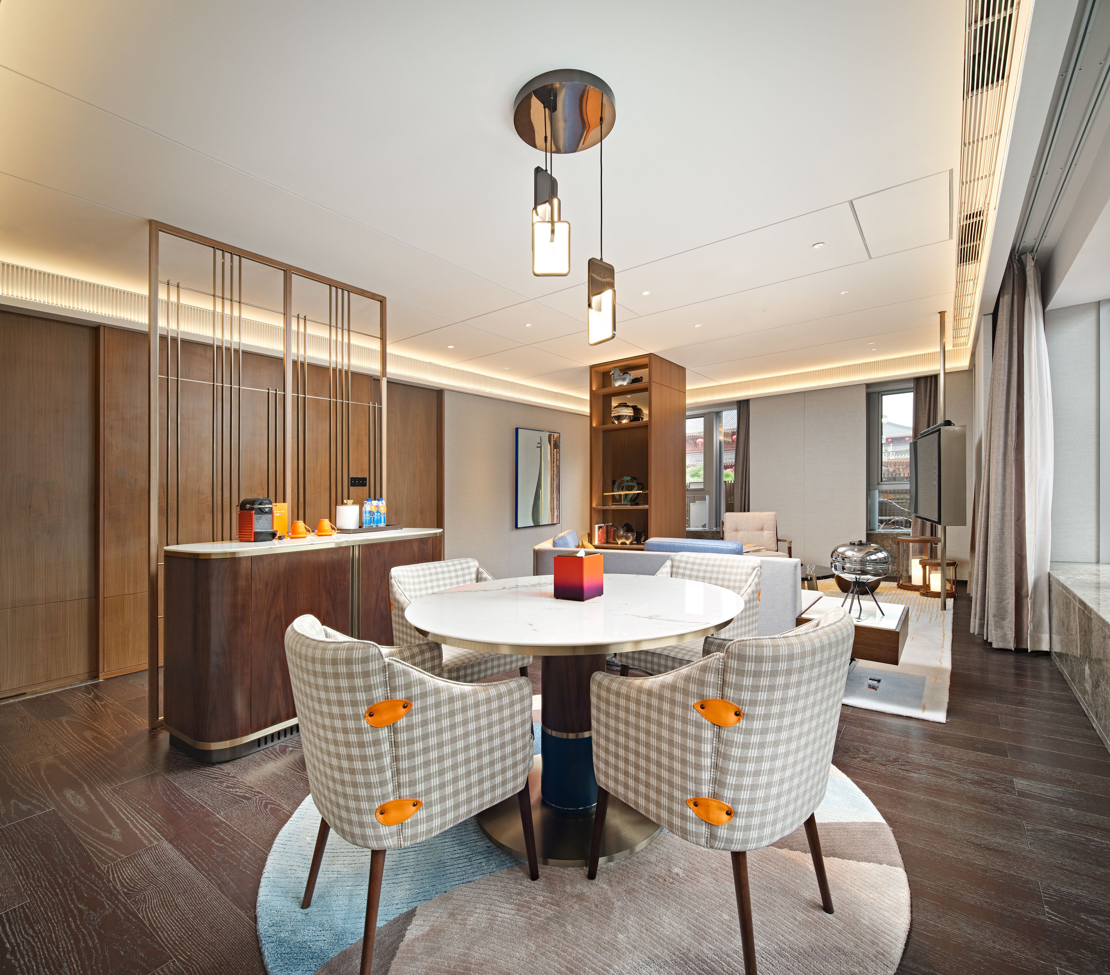 A modern hotel room with a round dining table, checkered chairs, a minibar, a large window, and a view of the city.