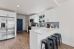 A modern kitchen with white cabinetry, a stainless steel refrigerator, a microwave, and a large island with bar stools. - Spacious, pet-friendly home near downtown Spokane—sleeps 14. (Spokane)