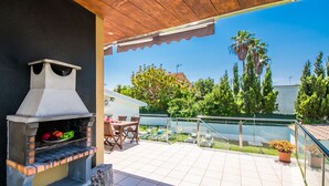 Een terras buiten met een barbecue, zithoek en uitzicht op de groene omgeving en gebouwen.