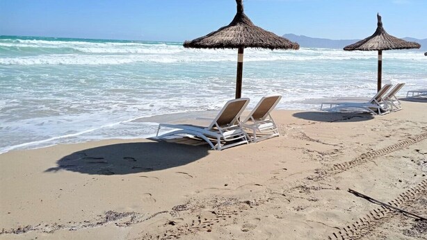 Een strand met twee rieten parasols en witte ligstoelen.