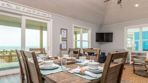 A dining area with a wooden table set for four, a television mounted on the wall, and a view of the ocean through large windows. - Sunny and 75 | August 1 opening. Pool, Gulf Front (Dauphin Island)