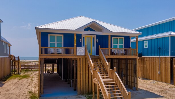 A two-story beach house with a wooden staircase leading to a balcony, situated on a sandy beach with a clear view of the ocean. - Sunny and 75 | August 1 opening. Pool, Gulf Front (Dauphin Island)