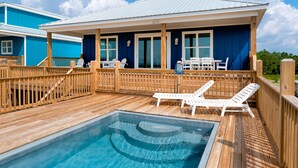 A wooden deck with a pool, lounge chairs, and a building with large windows. - Sunny and 75 | August 1 opening. Pool, Gulf Front (Dauphin Island)