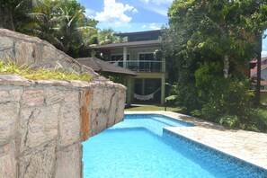 Una piscina con bordes de piedra, rodeada de exuberante vegetación y una casa de dos pisos con tejado de baldosas.