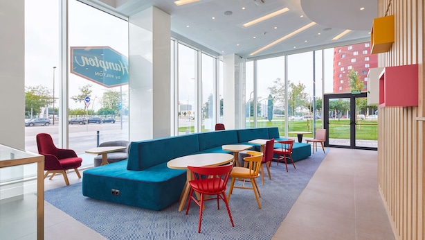 Un moderno vestíbulo de hotel con ventanas amplias, un sofá azul y sillas rojas y amarillas.