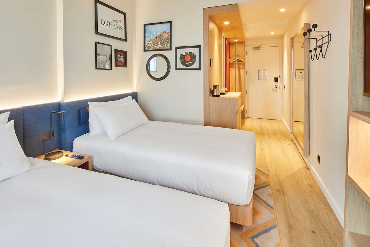 Een hotelkamer met twee bedden, een nachtkastje met lamp, een spiegel, ingelijste foto’s aan de muur en een houten vloer.