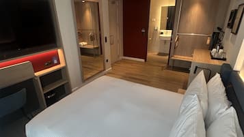 Una habitación de hotel moderna con una cama grande, un escritorio con televisor de pantalla plana, un espejo y un baño visible a través de una puerta abierta.