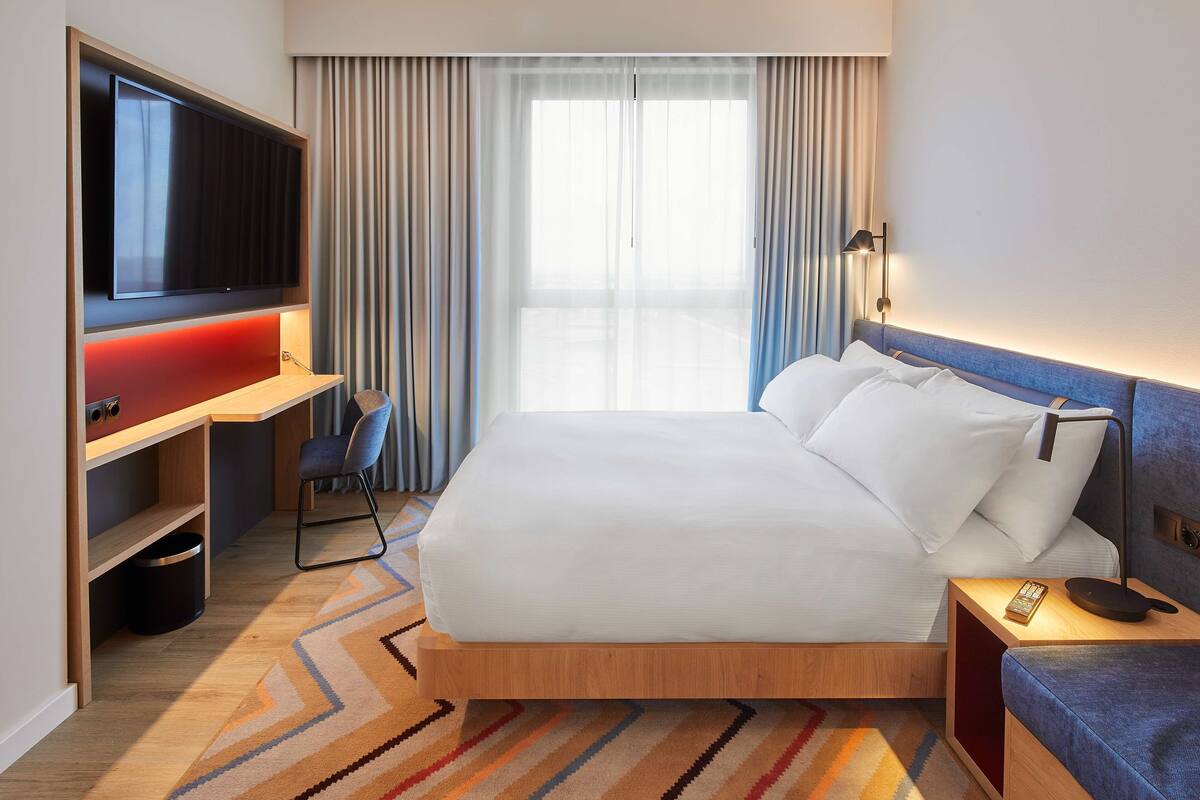 Hotelkamer met een bed, een bureau met televisie, een stoel, een nachtkastje en een raam met gordijnen.